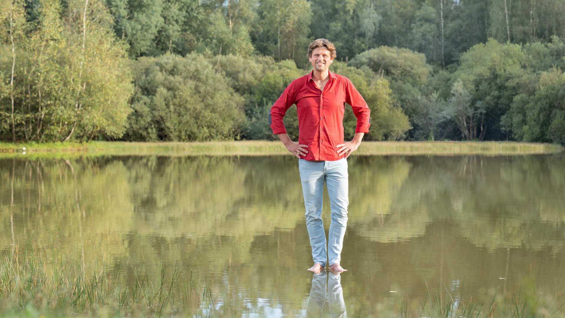 Alfred Slomp staat met blote voeten op een waterplas. Hij draagt een felrood overhemd, heeft zijn handen in zijn zij en kijkt de camera in.